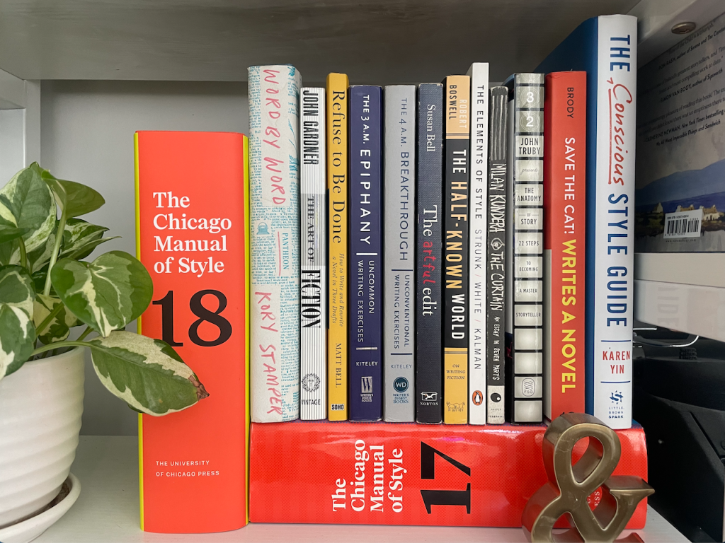 A stack of editing books sits of a shelf — including two editions of the Chicago Manual of Style, Word by Word, The Art of Fiction, Refuse to Be Done, The 3 A.M. Epiphany, The 4 A.M. Breakthrough, The artful edit, The Half-Known World, the Elements of Style, The Curtain, The Anatomy of Story, Save the Cat Writes a Novel, and the Conscious Style Guide. A potted plant sits to the left.